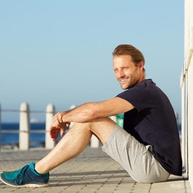 full body shot of an adult male sitting on a pier by a beach